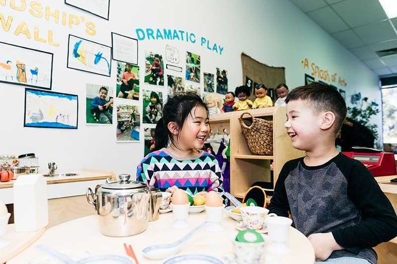 Two preschool children sit at a table set up for pretend play. On the table are a large silver tea pot, tea cups and saucers and breakfast items such as boiled eggs and fruit. The children are engaged and happy. In the background is a sign on the wall. The blue writing spells, Dramatic Play.