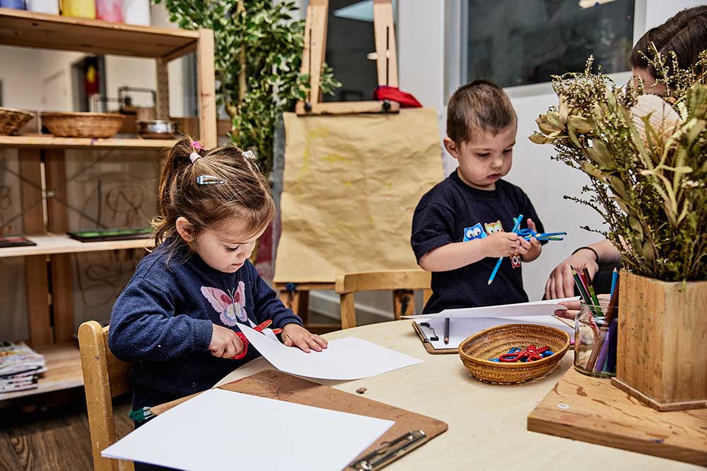 A child sits at a table in an early childhood education and care studio using a red pair of scissors to cut paper. There is a pretty embroidered butterfly on her jumper. Next to her stands a child holding a pair of scissors and a blue colouring pencil. On the table in front of them is a basket of colourful scissors and a glass bottle with colouring pencils. There is a clipboard with plain white paper on it. An educator sits at the table. Their face is partially blocked by Australian flowers. The educator holds out a piece of paper to the child. Behind them in the background is an art board with brown paper on it and wooden shelving wtih various loose parts and resources.