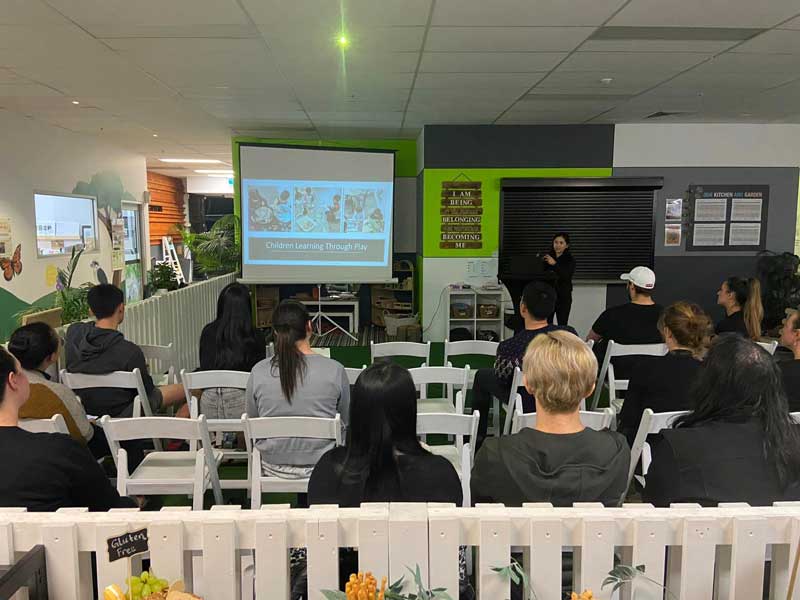 An educator stands in front of a audience communicating to parents at an evening event about children learning through play.