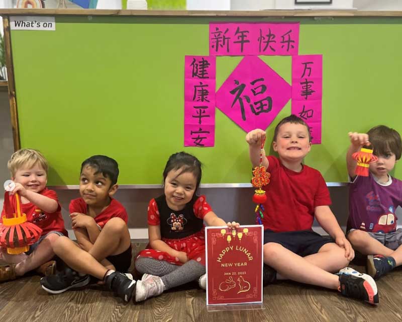 A group of children wearing shades of red and holding decorations sit in front of a Chinese lettering wishing everyone a happy Lunar New Year 