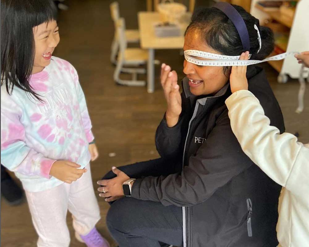 An educator is stting on a chair as two children use a tape measure to learn the size of her head as part of a inquiry-based learning project. One child watching is laughing while the other holds the tape up. The educator is also laughing with one hand just in front her head.