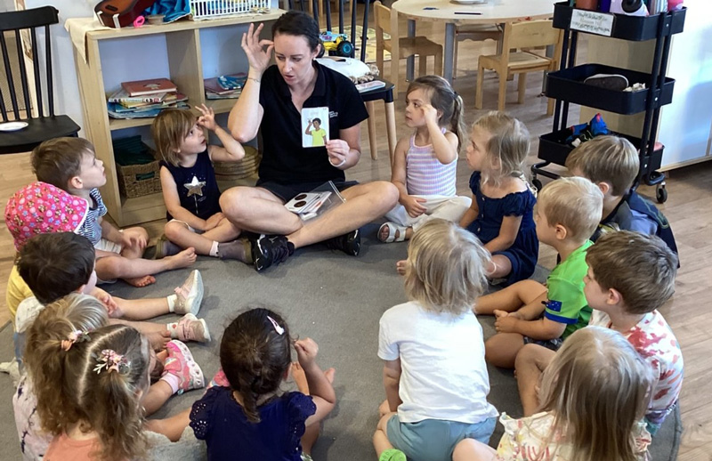 A group of children from Petit ELJ Burleigh sit around an Educator as the learn how sing using Auslan.