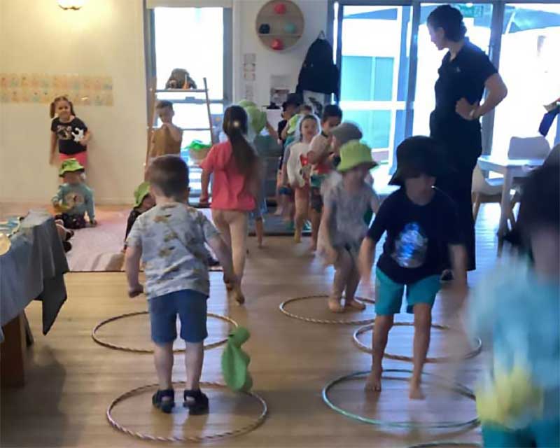 A group of preschool children bring an outdoor activity inside. On the floor are several hula hoops. Children are jumping from one hoop to the next.. There are two lines, one group of children are jumping forward towards the camera and the other are jumping away with their backs turned. An educator supervises the indoor game and a other children at the back of the room wait their turn.