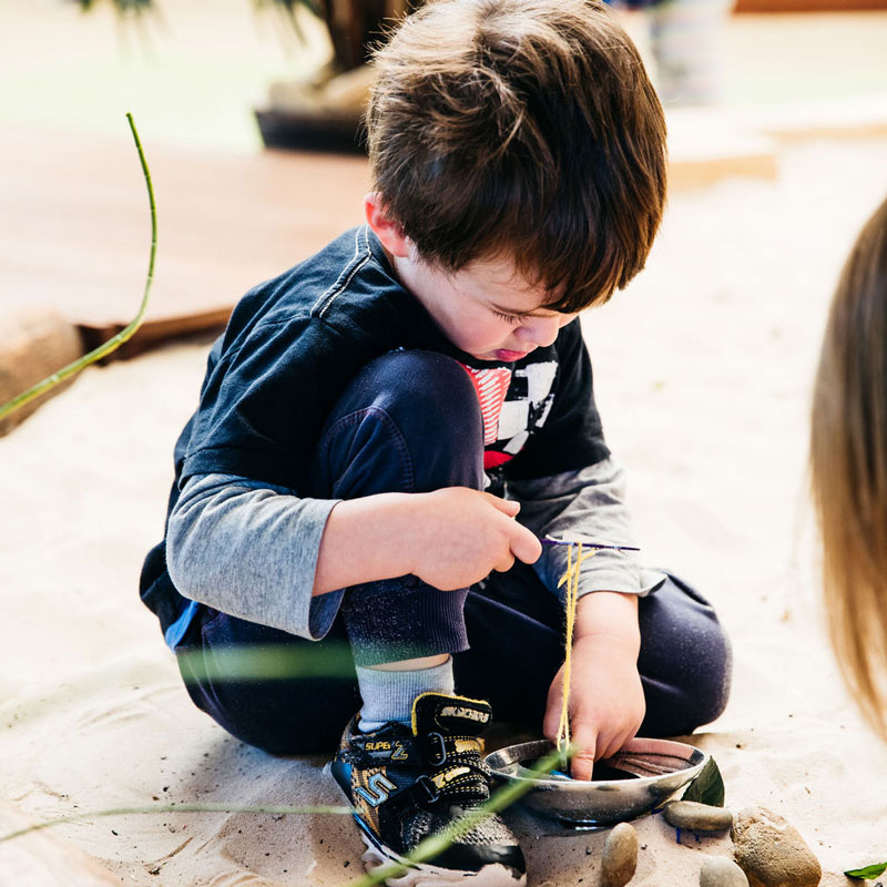 A child sits in a sandpit playing with loose parts including kitchenware, rocks and grass. Another child sits near by, their long hair partially noticeable Big life changes includes transitions and continuing routines such as play.
