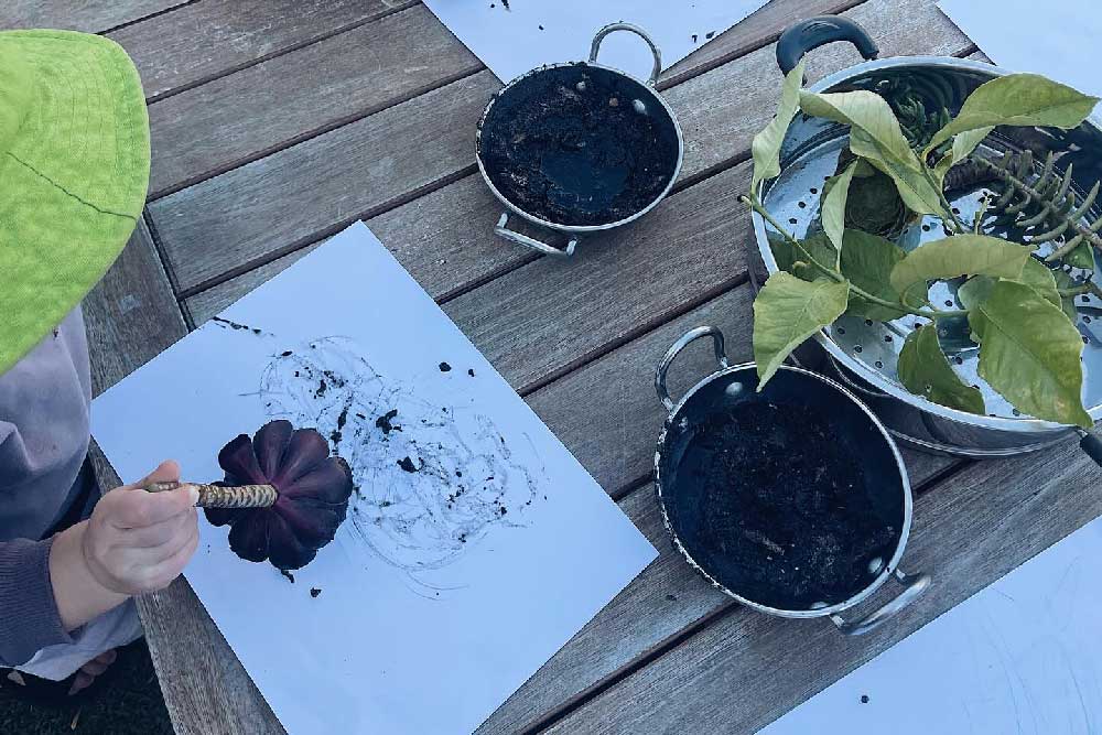 A child sits at an outdoor table participating in art. They hold a large red flower in one hand. They are creating an art piece using the recycled flower as their brush. In front of them are two large pots filled with mud. The mud is their paint. Behind the the two pots is a larger steamer pot with branches and leaves.