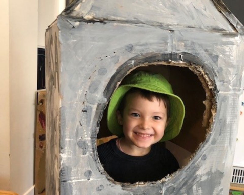 A pre-schooler stands inside a spaceship made from painted cardboard for science week encouraging children's imagination for play and learning. 