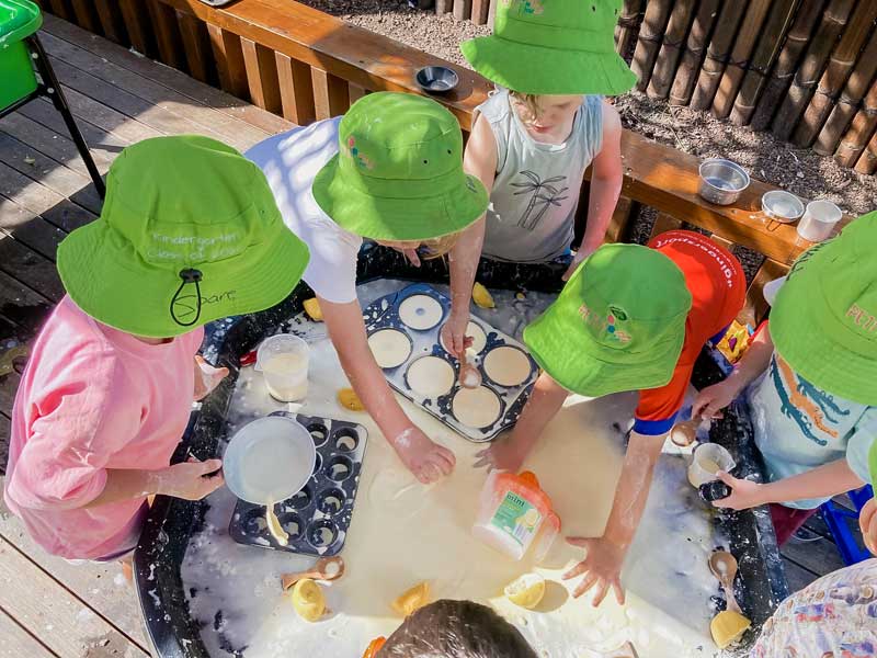 A group of children gather around a sensory table filled with goop and slices of lemons in an outdoor area. Some children are filling baking dishes with the slime, while others are getting messy with their hands.