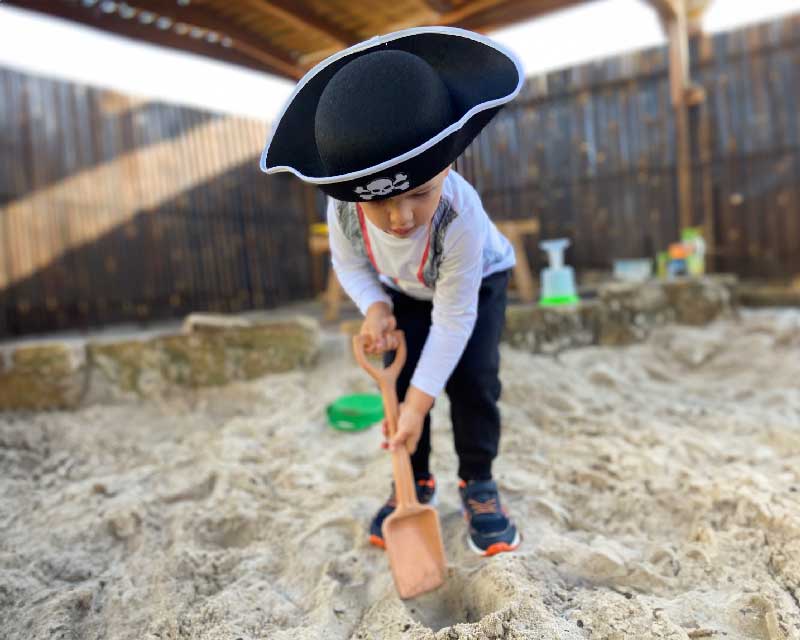 A child wearing a pirates costume and holding a cream coloured shovel digs deep into the sand. Behind him is a wooden fence and over a wooden pergola.