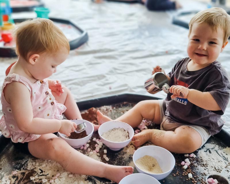 Two children face each other in a sensory tub where they are exploring messy play with cocoa powder, flour and marshmallow. Sensory experiences are fun stocking stuffers ideas for children.