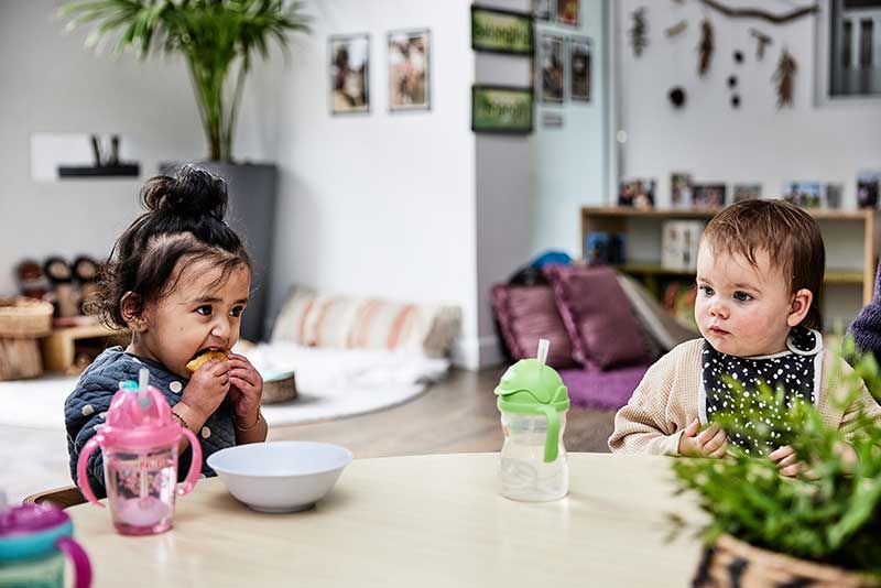 Two children sit at a round wooden table. One child has a white bowl and has both hands around food that they are eating. Both children have colourful water bottles. The child eating has a pink sipper cup water bottle and the other child has a green sipper cup water bottle to encourage them to drink water. 