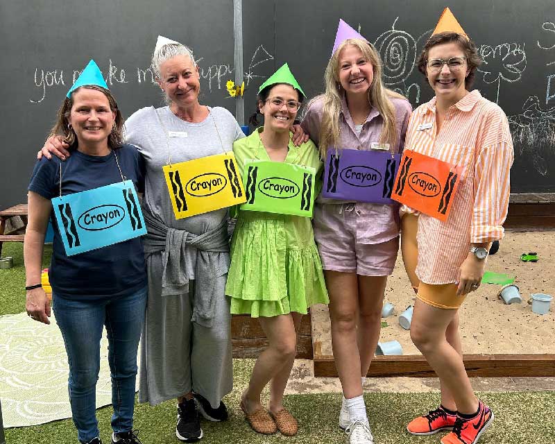Five educators from Petit Early Learning Journey stand together, linking arms as they pose for children's book week as colourful crayons. Behind them is a sand pit and a chalk board. 