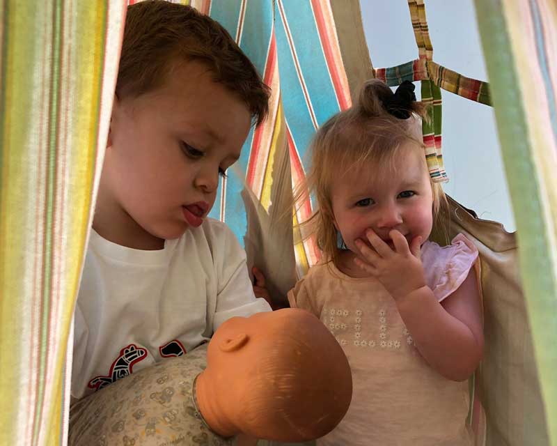 Two children pretend they are parents of a child. They play in a tent which is a home and the boy holds a baby doll. Improvising stories and roleplaying is a fun play-based tool.