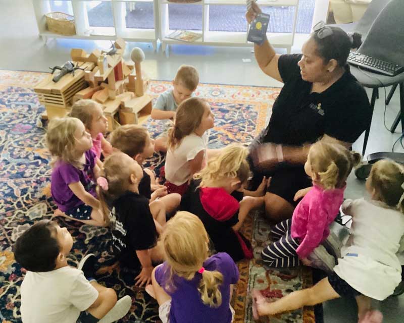 Children gather around sitting on a mat in an indoor setting with Acting Assistant Centre Director and Educational Lead Letitia Da Silva from Petit ELJ Marian, holding up a small book.