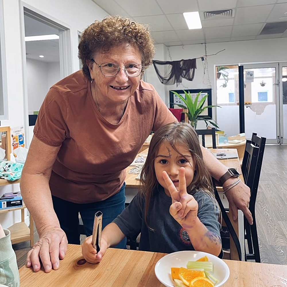 A grandmother stands behind a child inside a Petit Early Learning Journey studo. The child is sitting at a table. On the table in front of them is a bowl of fruit. The child is making the peace sign in their left hand and holding a pair of tongs in their right. The child has paint on their left hand and on their face. Behind the grandmother and child is a table and corner set up with learning and play experiences.