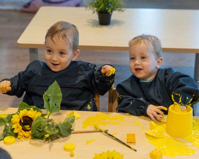 Two children sit behind a desk enjoying a sensory activity involving sunflowers, yellow paint, round and square yellow blocks.