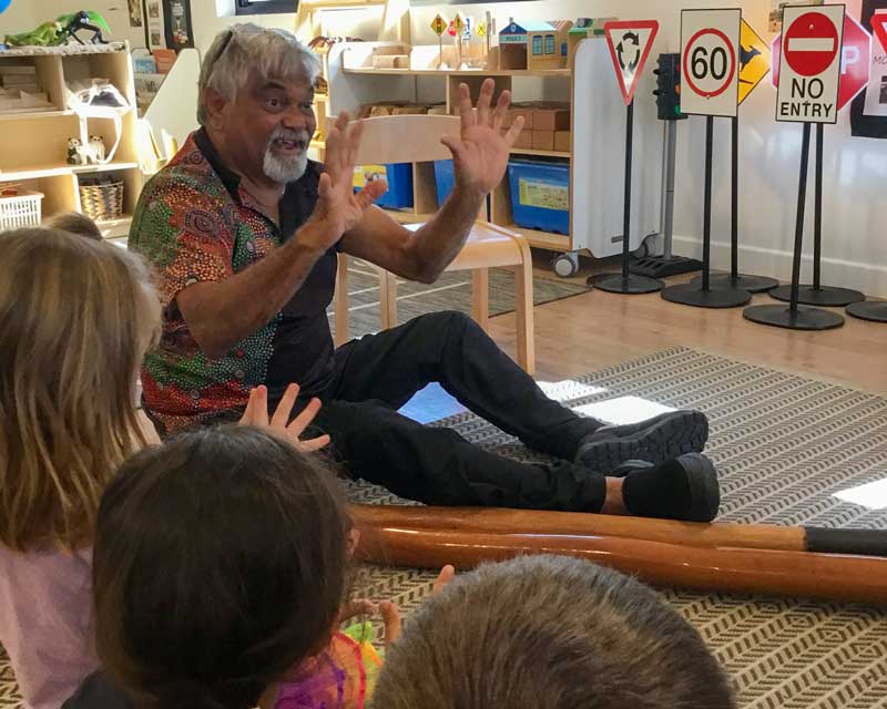 Uncle Alan, a local Aboriginal Elder sits on the floor with a didgeridoo at his feet. He teaches children about the land we are on. He shares his knowledge through the ancient art of oral storytelling.