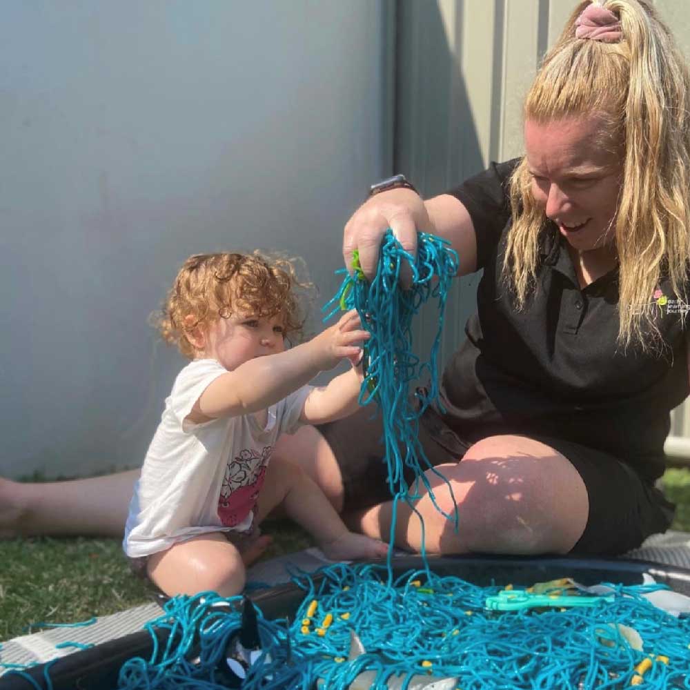 An educator sits on the ground next to a child. She holds up a handful of colourful wet spaghetti from a sensory bin as the toddler reaches towards it. She observes and listens to the child building relationships and trust.