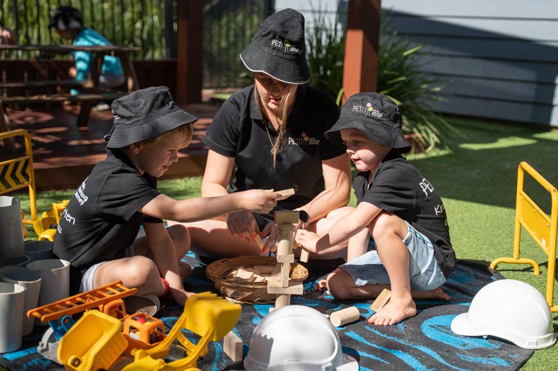 Two children and an educator sit outdoors on a blanket with homemade wooden blocks, ideal children's stocking stuffers. They are building a tower together. Other resources are around them including construction signs, toy trucks, short pipes and construction hats.