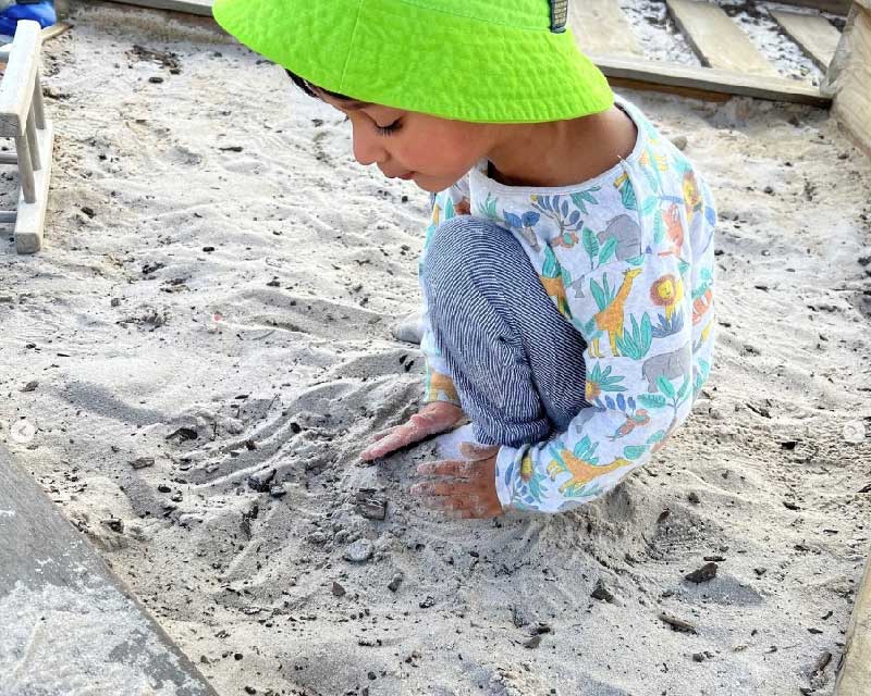 A child wearing a green hat and a long sleeved shirt animal patterned shirt and stripped blue pants crouches down in the sand building a hill and benefitting from sandpit play.