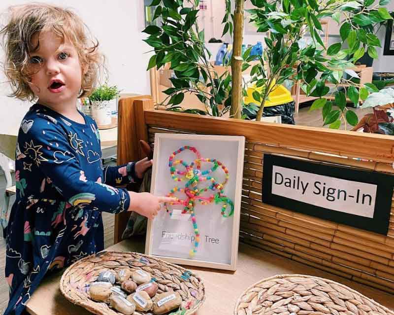 A child stands beside a daily sign-in desk that uses stones with children's names on them. The child points to a friendship tree made of beads in an enclosed glass frame. A visual display, the friendship tree is a form of documentation that remind children of teamwork.