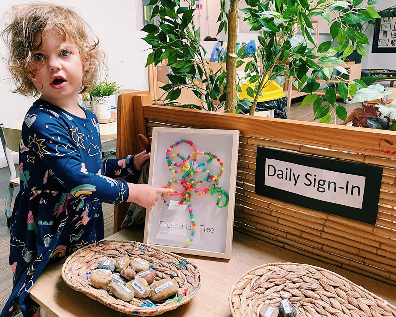 Child points to a group framed art piece "Friendship Tree" at a sign in table enacting rituals with their families and Educators for a smooth drop off - an example of the Circle of Security in action.