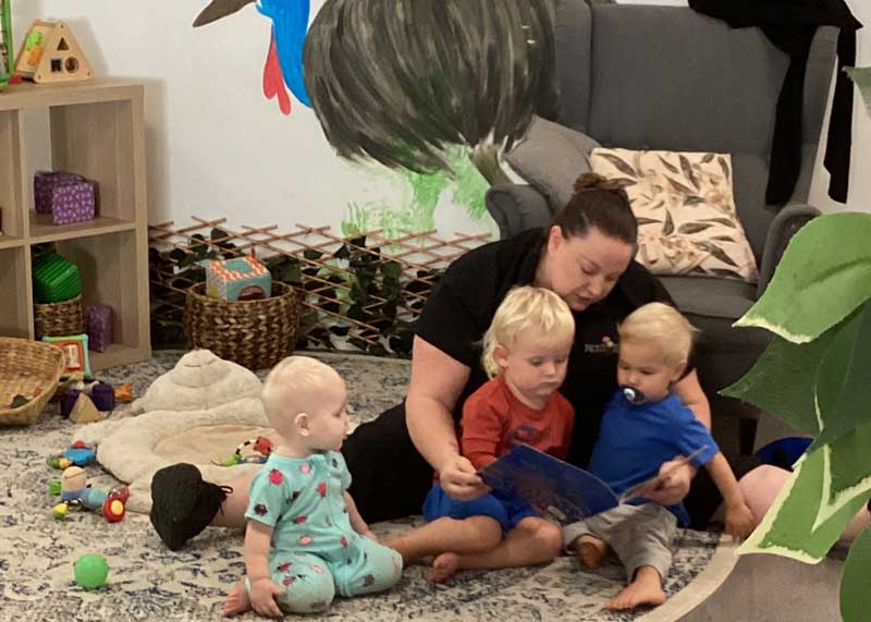 An educator from Petit ELJ  shares an appreciation for Indigenous culture by reading Colours of Australia by Bronwyn Bancroft. Three children sit in front of the educator who sits behind them on the floor. In the background is a large chair and a painting of a cassowary.