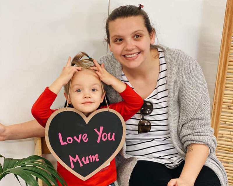 Child with sign that reads "Love You Mum" sitting next to their mum for Mother's Day.