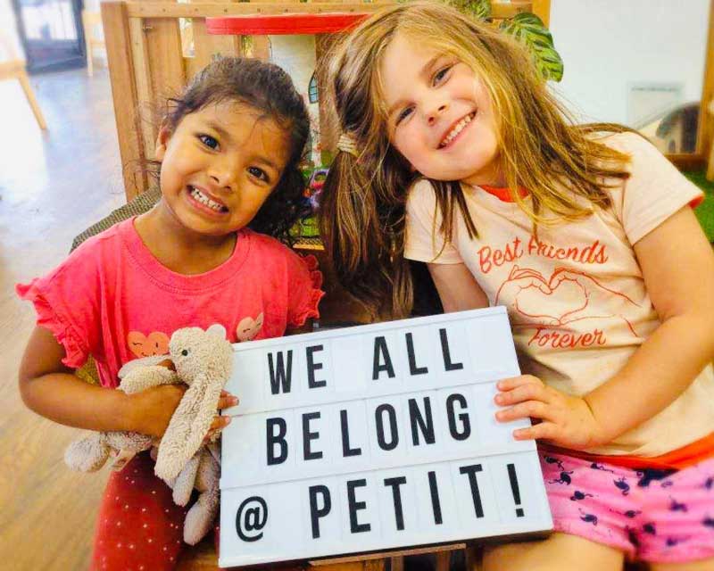 Two children hold a sign between them that reads "We all belong at Petit". One child also holds a couple of rabbit plushies. Both children are smiling. A painting easel sits to one side in the background. 