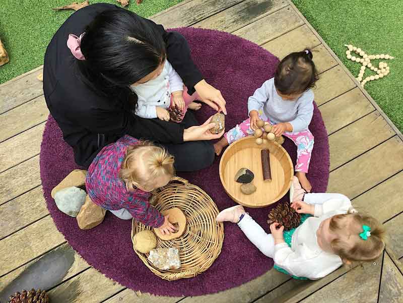 An educator makes children's learning visible by co-constructing learning on a purple round mat on top of a wooden porch. A child sits on her lap and three other childre sit around the mat. They are all playing with natural resources taken from a basket and wooden bowl. They include rocks, pine cones, seeds and a branch.