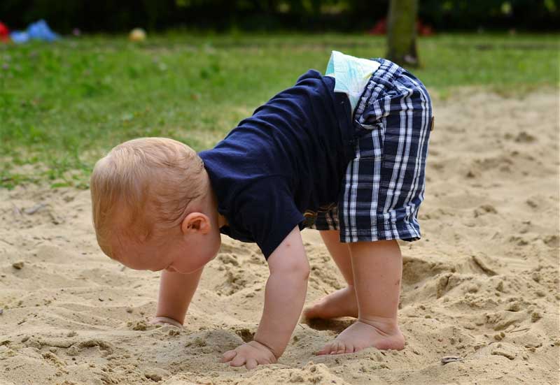 A child bends over touching the sand with their nappies visible under their shorts. To wear cloth vs disposable nappies is a question many families face.