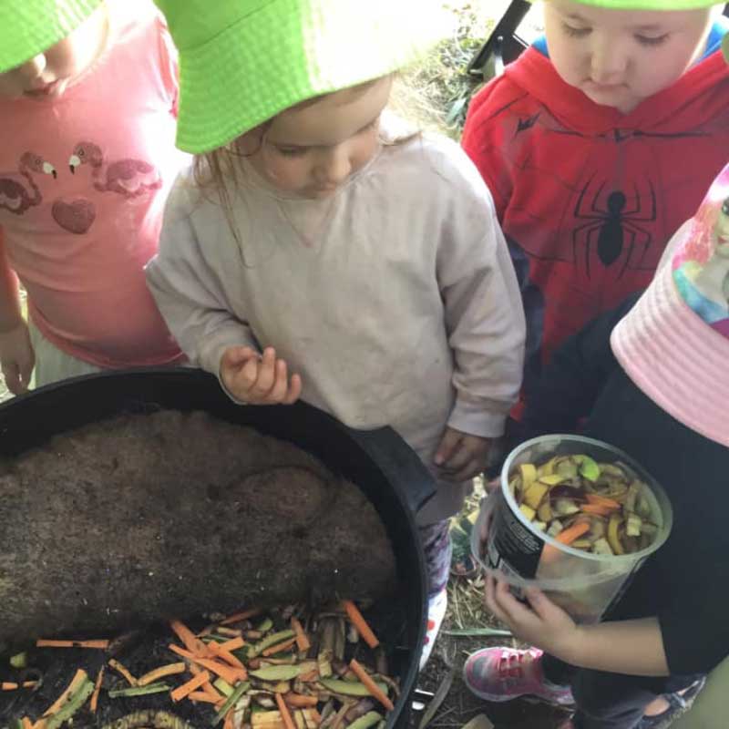 ALT: Children gather around a compost bin filled with unwanted fruit and vegetables. One child holds a bucket with scraps.