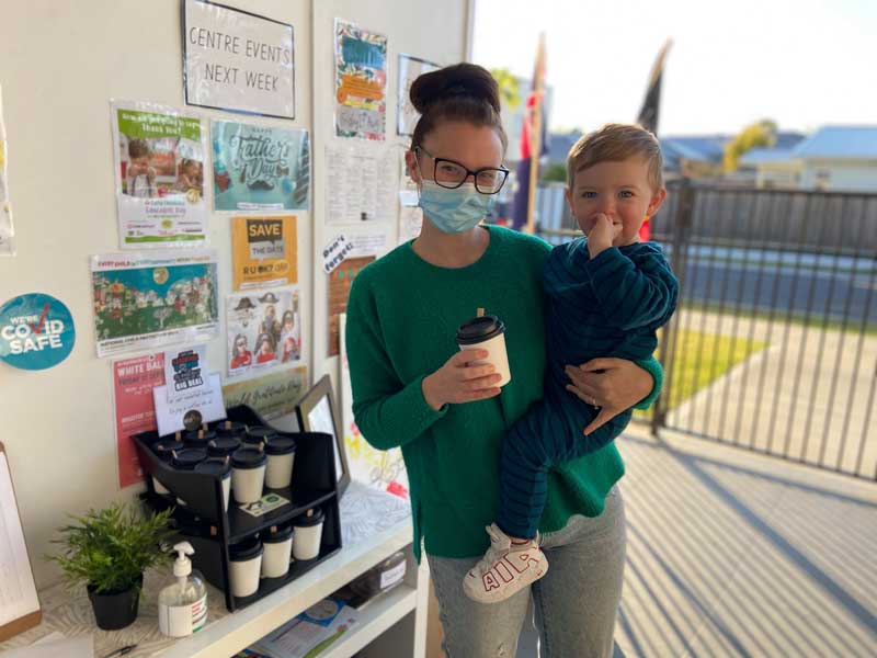 Mum holding a child stands in front of a noticeboard with a sign reading "Centre Events Next Week", one of the different ways of communicating with parents.