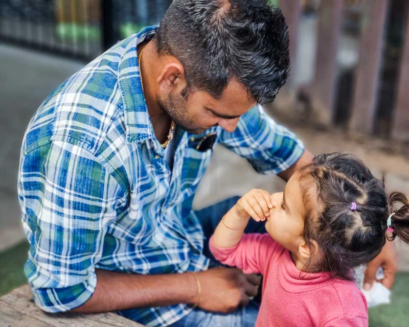 A man sitting next to a wooden table looks down into the eyes of his daughter while celebrating Father's Day with other families.