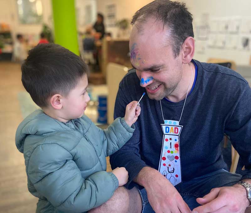 A child's special person honours Father's Day traditions by sitting on a small stool so a child holding a cotton tip painting the man's face blue with makeup. The man is wearing a handcrafted tie.