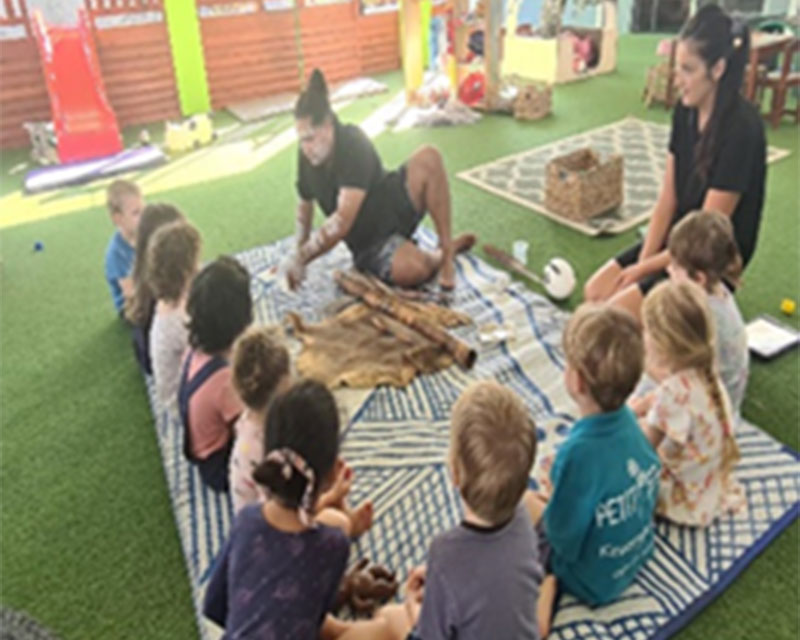 Children sit on a mat in an outdoor area at Petit ELJ. They are listening to a local indigenous family member with arms and face covered in ochre body paint share indigenous perspectives and stories. On the mat is a didgeridoo and kangaroo animal hide.