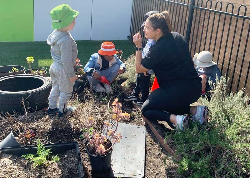 An educator kneels on a garden bed surrounded by children as she listens and provides indoor and outdoor gardening tips.