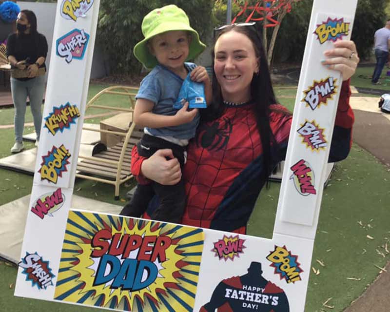 A special person and role model in a spiderman costume holds a boy in a green hat. They are holding a sign, "Super Dad" and celebrating Father's Day.