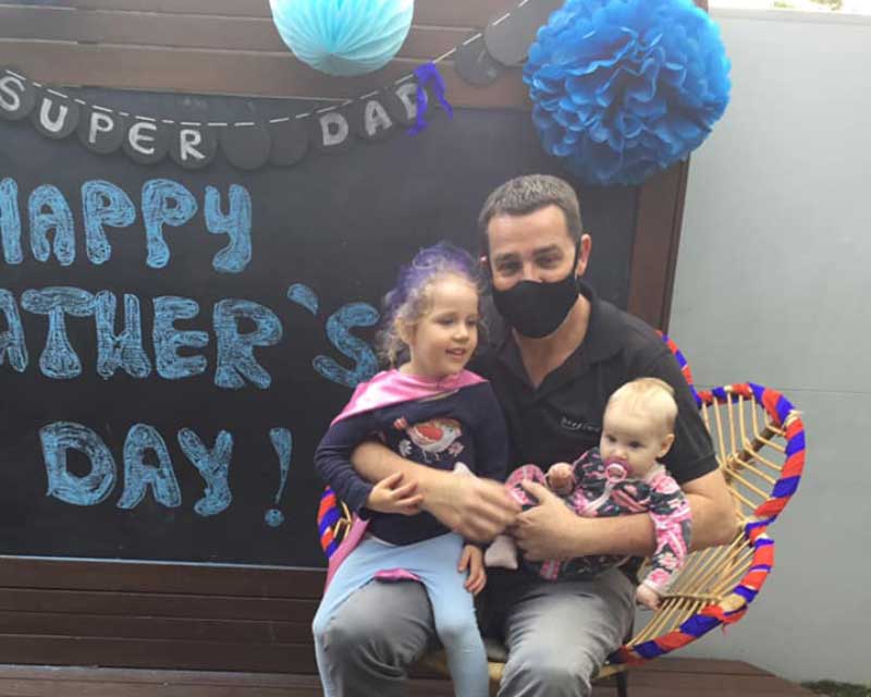 A dear Dad sits in a wicker chair holding two children in his lap, a preschooler and a toddler. They sit in front of a display with decorations that say, "Super Dad" and "Happy Father's Day".