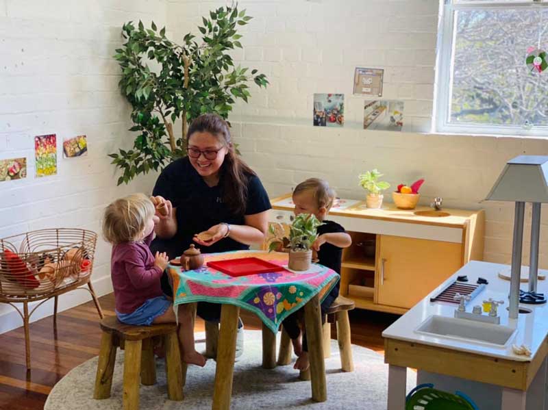 Educator sits at table between two children roleplaying and chatting and helping with language development. A tea service. to one side is a wooden cabinet and in front a make believe kitchen.