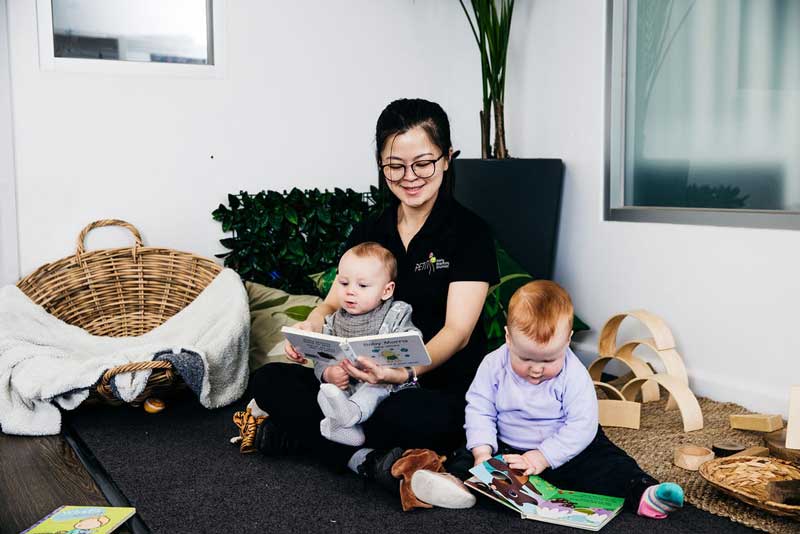 An educator reads to two young children encouraging positive learning dispositions. One toddler sits in the carer's lap, following the story. The other sits next to the educator and turns pages on a different book. the story while another.