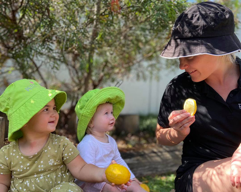 Two toddlers wearing hats. One holds a lemon. An educator sits with them tasting a slice of lemon.