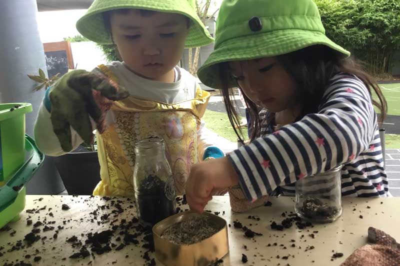 Two children play and learn while gardening as they stand at an outdoor table filling bottles with dirt and seeds to make terrariums. One child wears a gardening glove. There is lots of dirt on the table and a medium sized green bucket. In the background is an outdoor area with bike track.