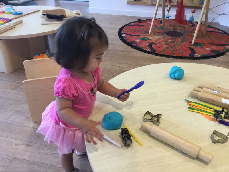 A child stands at a table with a blue ball of homemade playdough. She investigates the tools available.