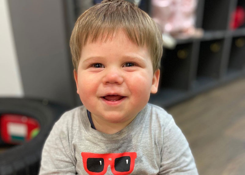 A child smiles at camera showing his first baby teeth. He wears a grey shirt with a sunglasses print. In the background is a tire and multiple cubby holes for belongings in an early learning service.