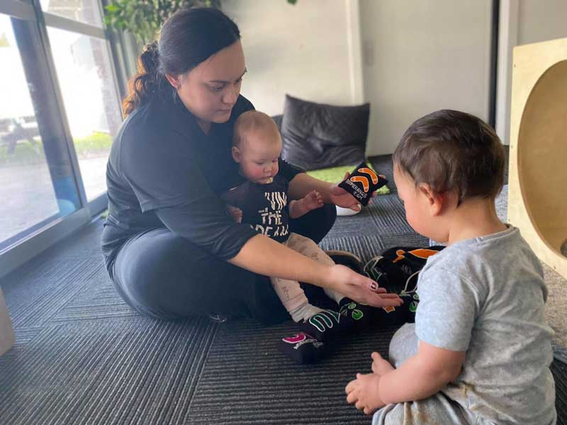 An educator from Petit ELJ Coffs Harbour sits with a child in their lap and another child across from them. Between them are hand-sized pillows decorated with symbols from local indigenous knowledge.