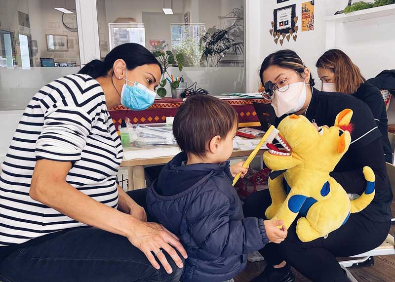 A child stands between two sitting educators wearing masks and promoting oral health. One educator holds a large lion plushie with big white teeth. The child brushes the plushie's teeth with an oversized toothbrush. 