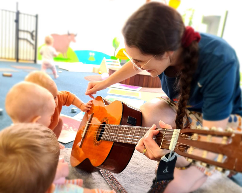 Three babies show their curiosity for sound and an acoustic guitar held by Katherine in our music program for babies at Petit ELJ Wooloowin.