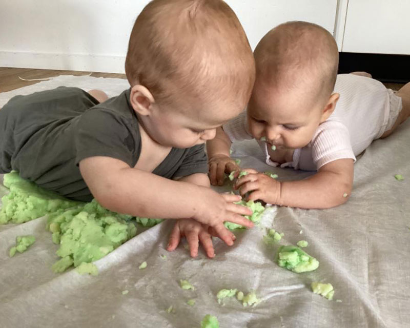 Two newborn babies share tummy time and a sensory experience with green edible playdoh on a white blanket. 