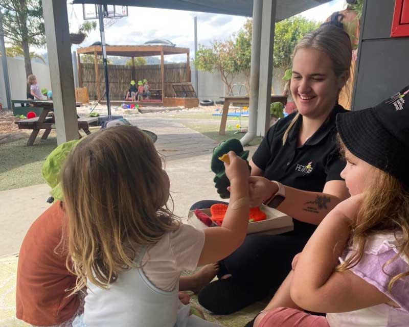 An Educator sits with children modelling good behaviour by showing her interest in their favourite book.