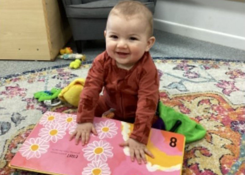 ALT: A baby with red cheeks wearing a brown all-in-one sits up on a round rug on the floor leaning over a big book with both hands flat on the pages. On the child's left is a white ring with a green toy. Cool teething rings can help soothe breastfeeding babies.